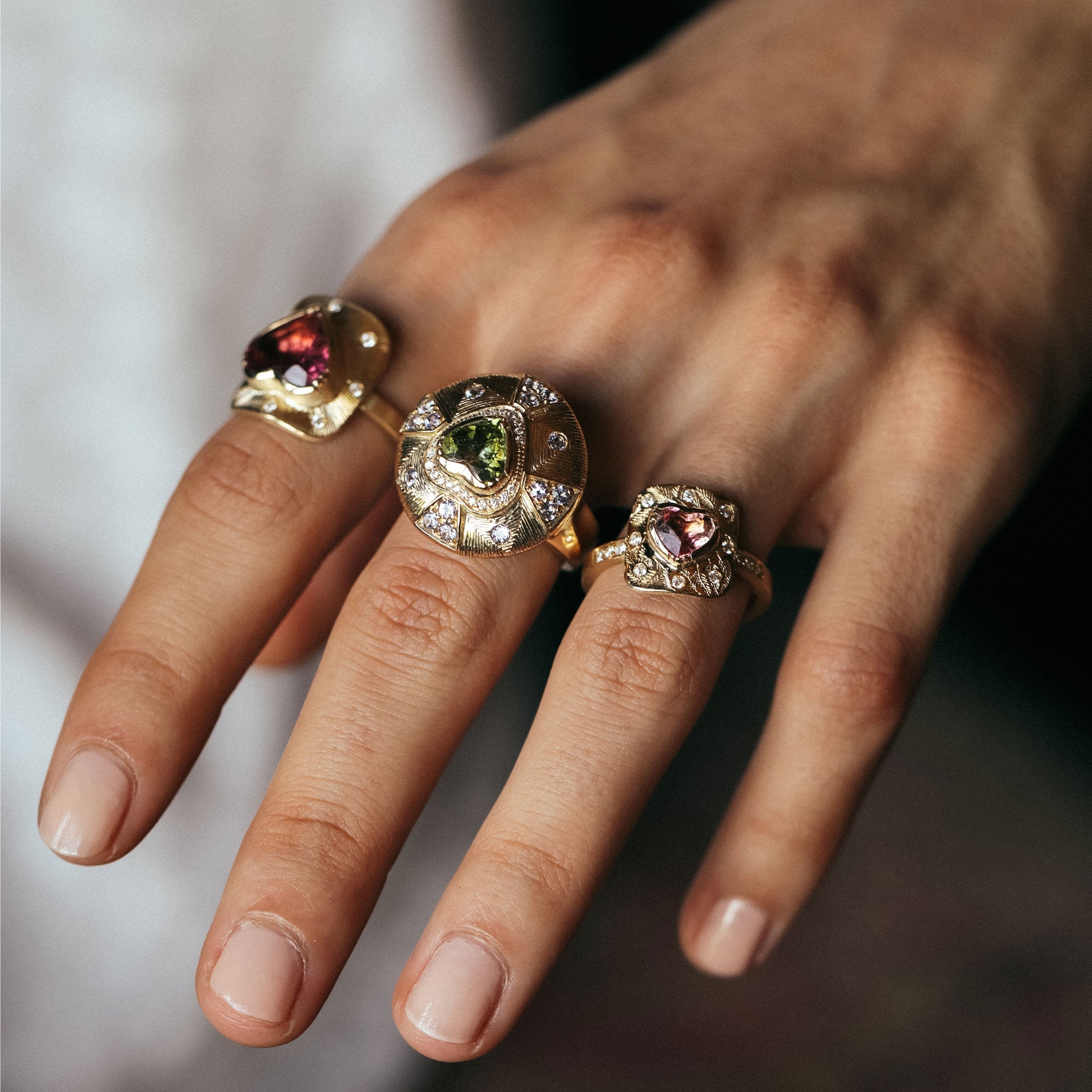 Hand wearing three gold rings with gemstones on a neutral background