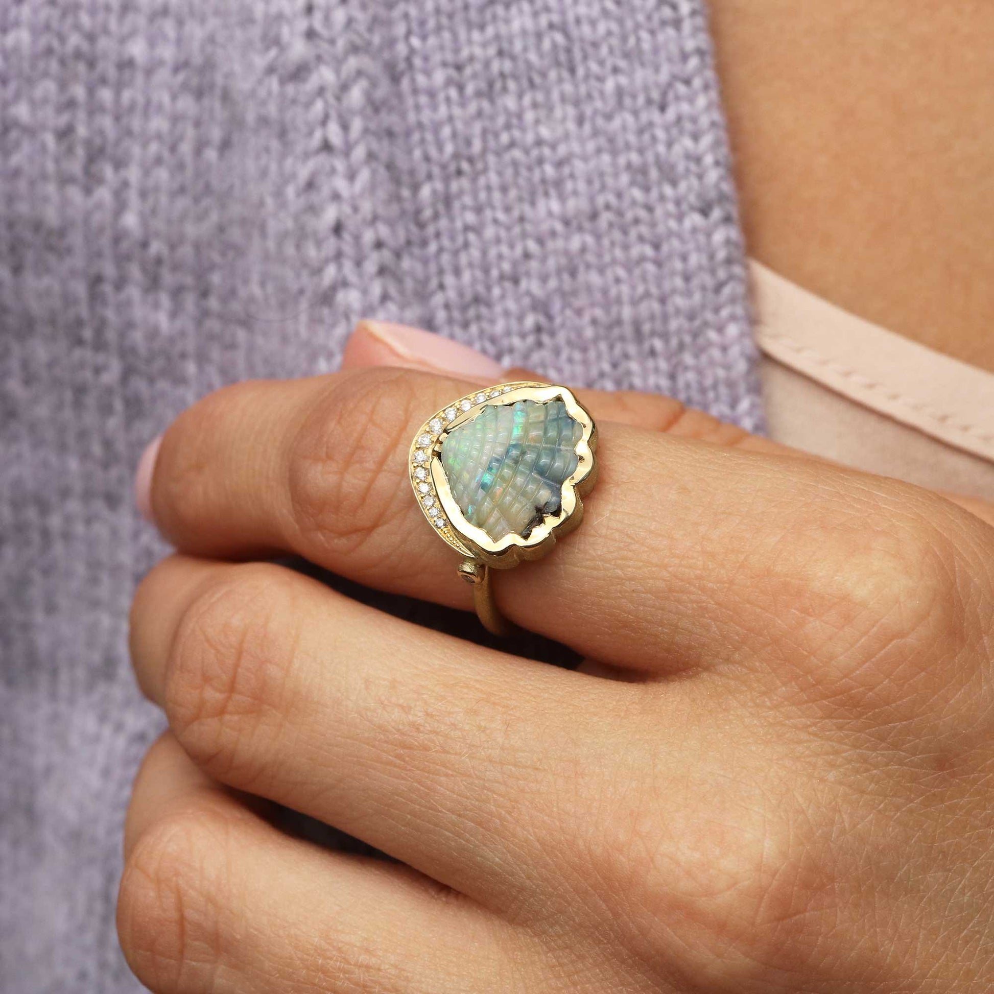A gold ring with a scalloped iridescent shell-shaped opal center stone and small diamond stones along the edge, worn on a finger. The background shows a person's hand, a light pink bra strap, and a textured lavender knit sweater.