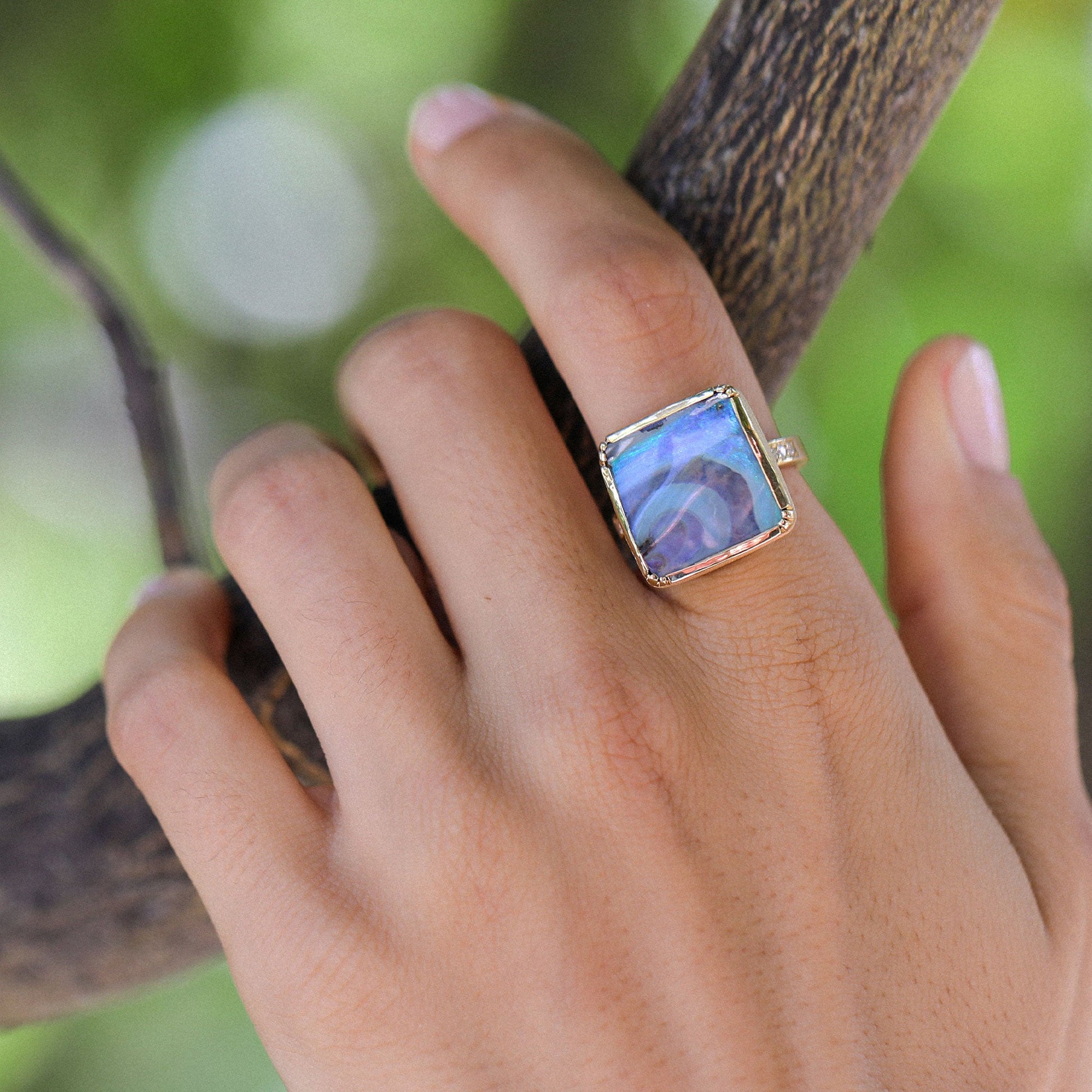 A close-up of a hand wearing a gold ring with a square iridescent blue-purple opal stone, set against a blurred green outdoor background.