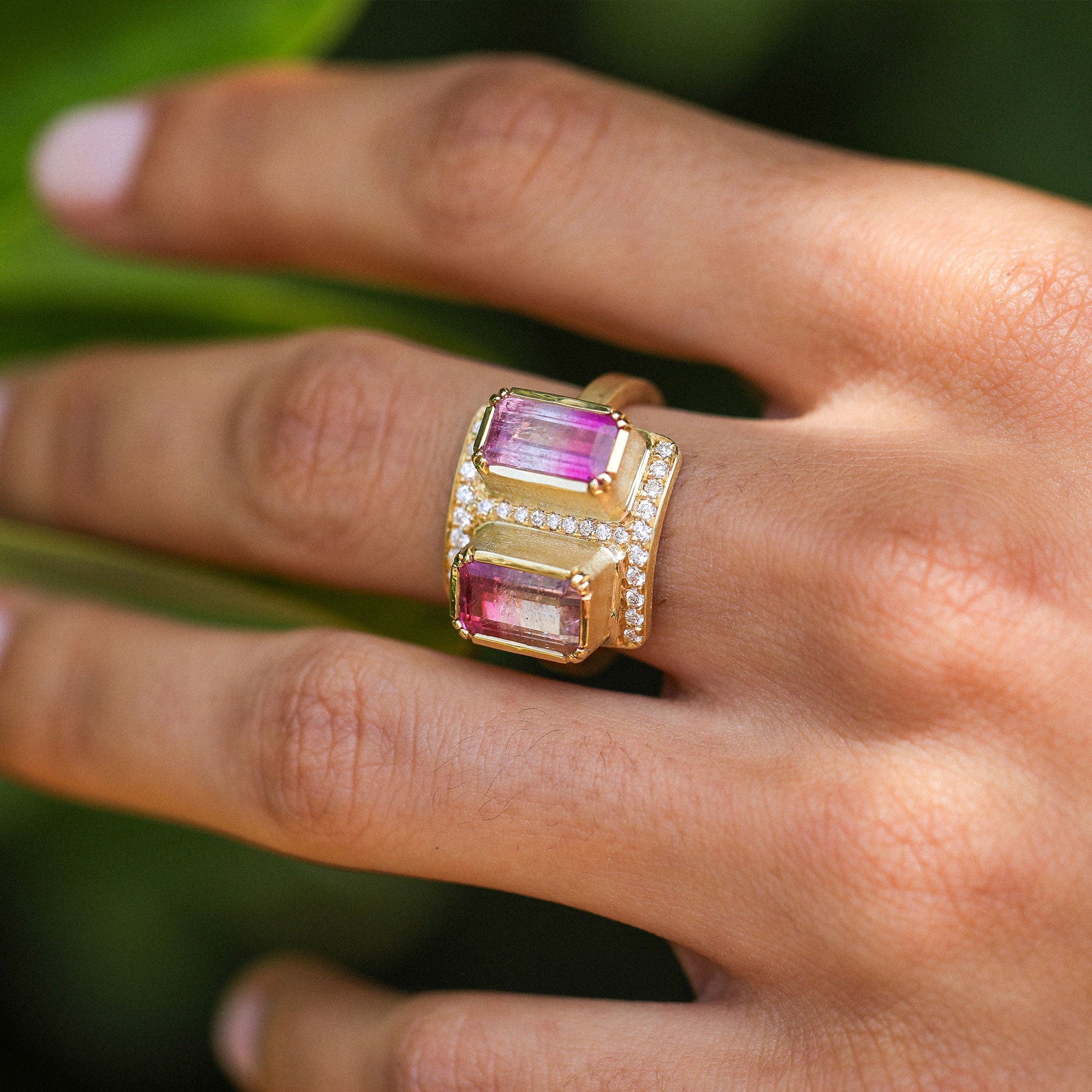 A close-up of a hand wearing a wide yellow gold ring with two rectangular pink bicolour tourmaline gemstones and rows of small diamond stones.