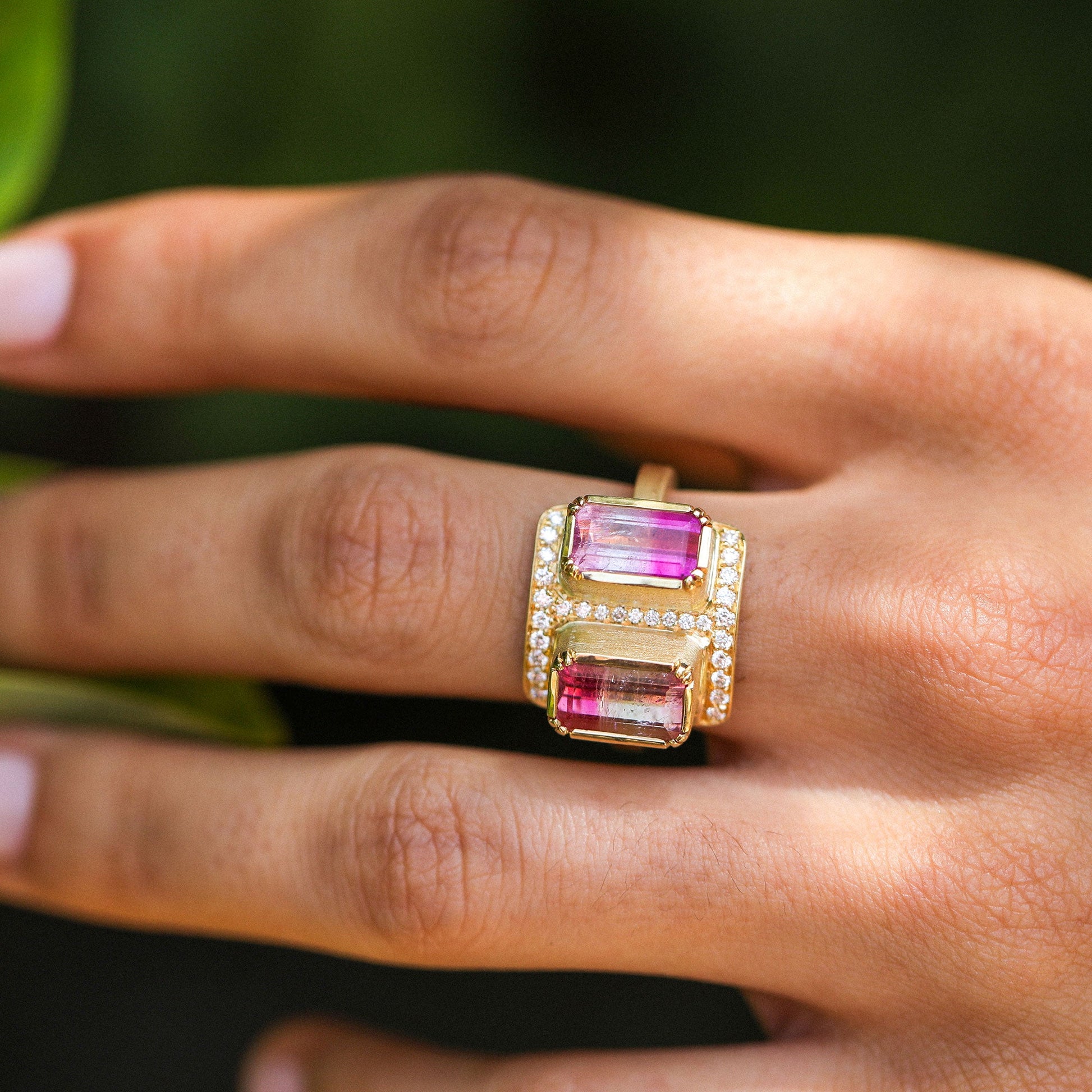 A close-up of a hand wearing a gold ring with two rectangular pink gemstones and small clear accent stones, set against a blurred green background.