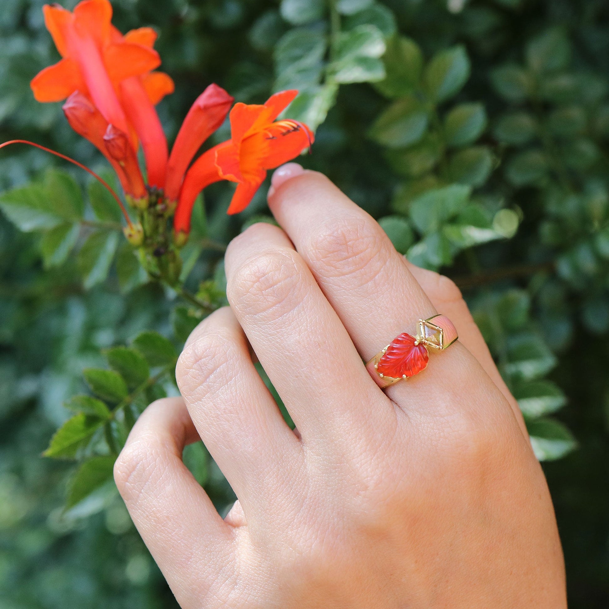Hand wearing a red fire opal and gold ring with a floral design, holding a red flower against a green leafy background.