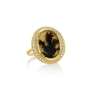 An 18k gold ring featuring an oval dendritic agate stone with a dark brown to black dendritic pattern, surrounded by a halo of small white diamond stones, set against a plain white background with a soft reflection.
