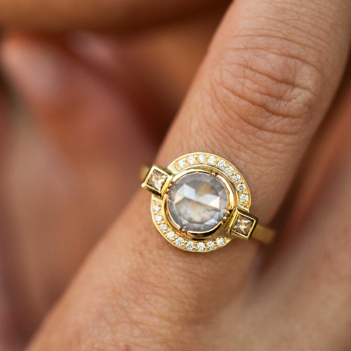 A close-up of a gold ring with a large round raw diamond rosecut gemstone and smaller accent white diamond pave stones, worn on a finger.
