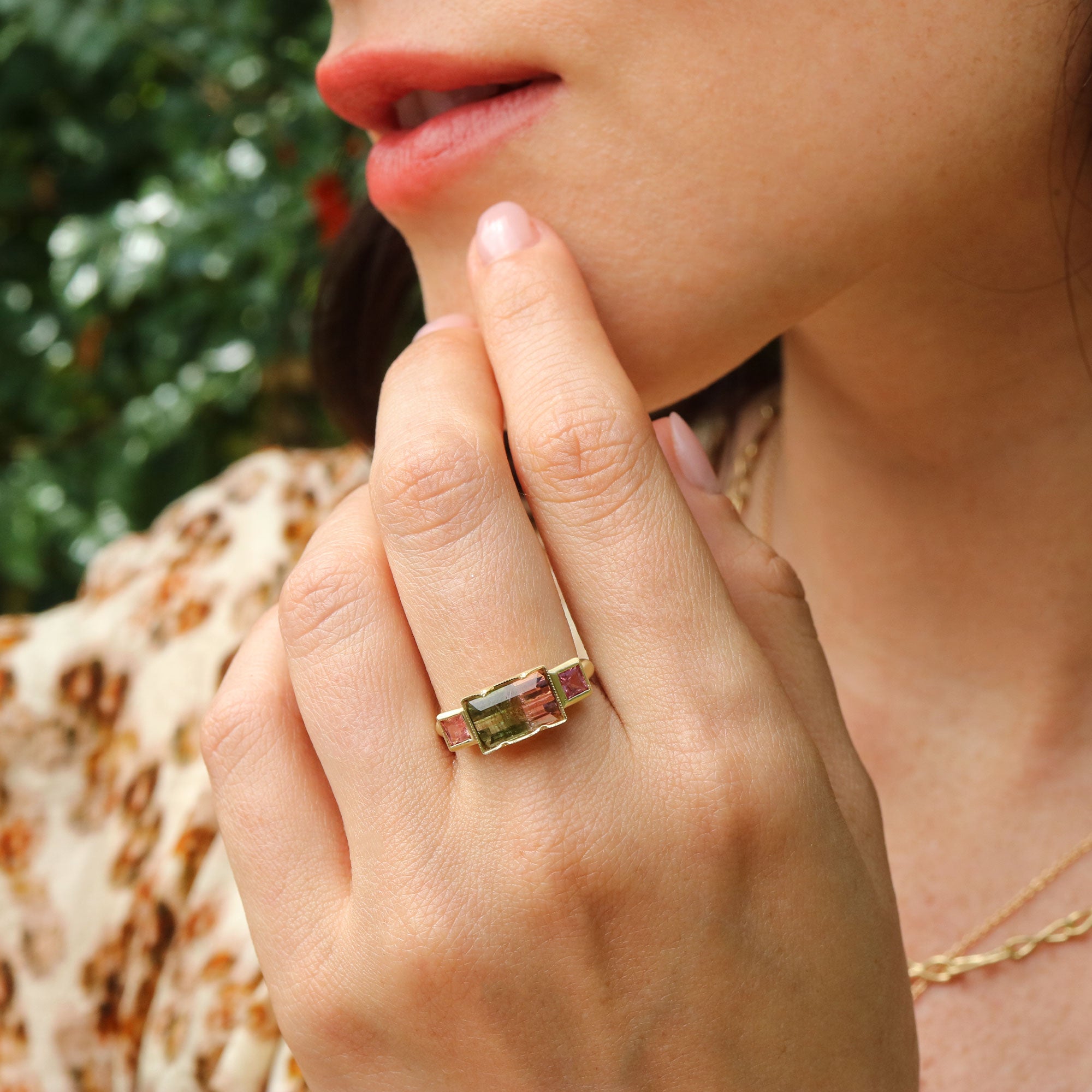 Close-up of a hand wearing a gold ring with a bicolor tourmaline gemstone and pink sapphires, outdoors.