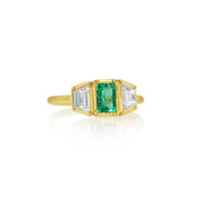 An 18k yellow gold ring featuring a central emerald-cut green emerald gemstone flanked by two baguette-cut diamond stones, set against a clean white background with a soft reflection.