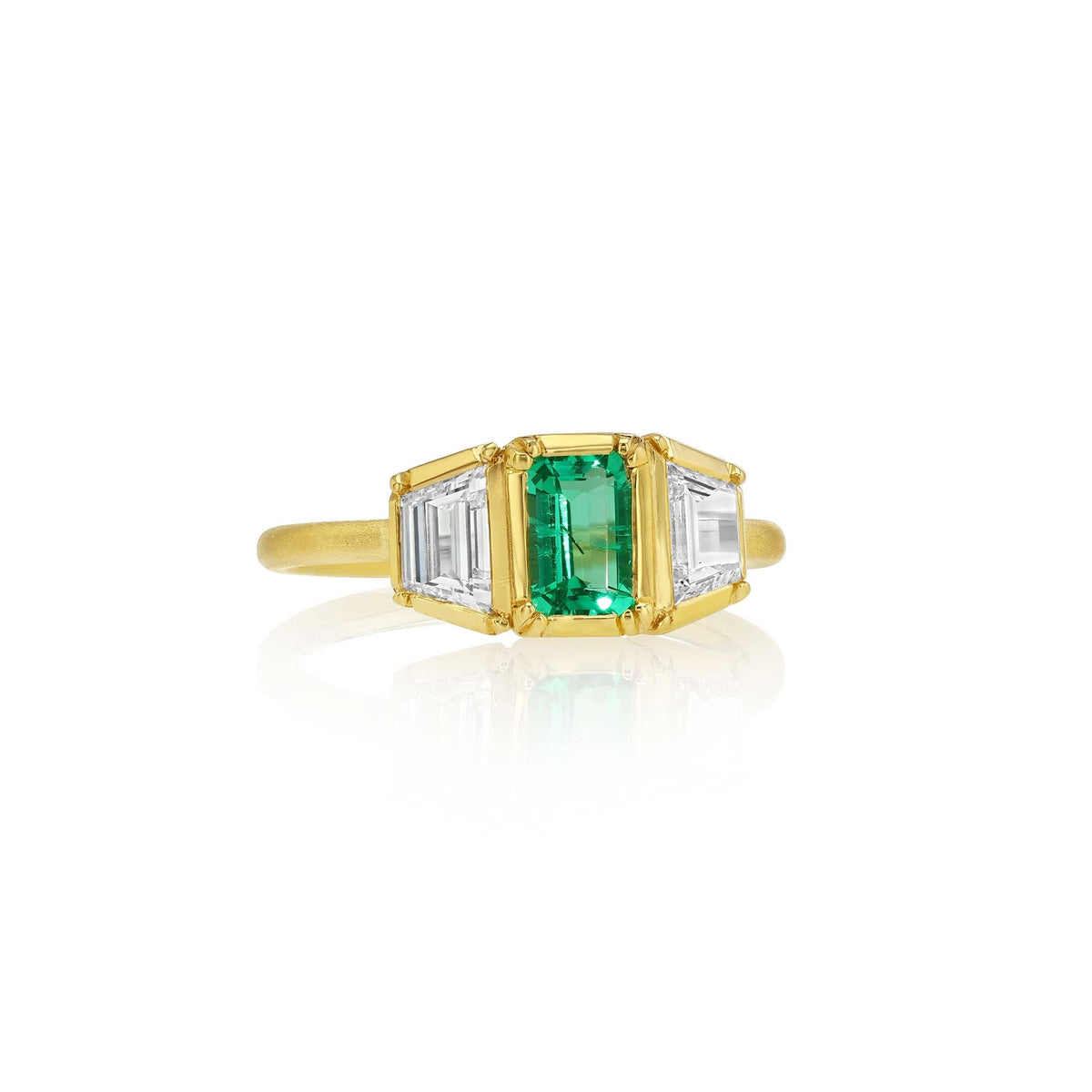 An 18k yellow gold ring featuring a central emerald-cut green emerald gemstone flanked by two baguette-cut diamond stones, set against a clean white background with a soft reflection.