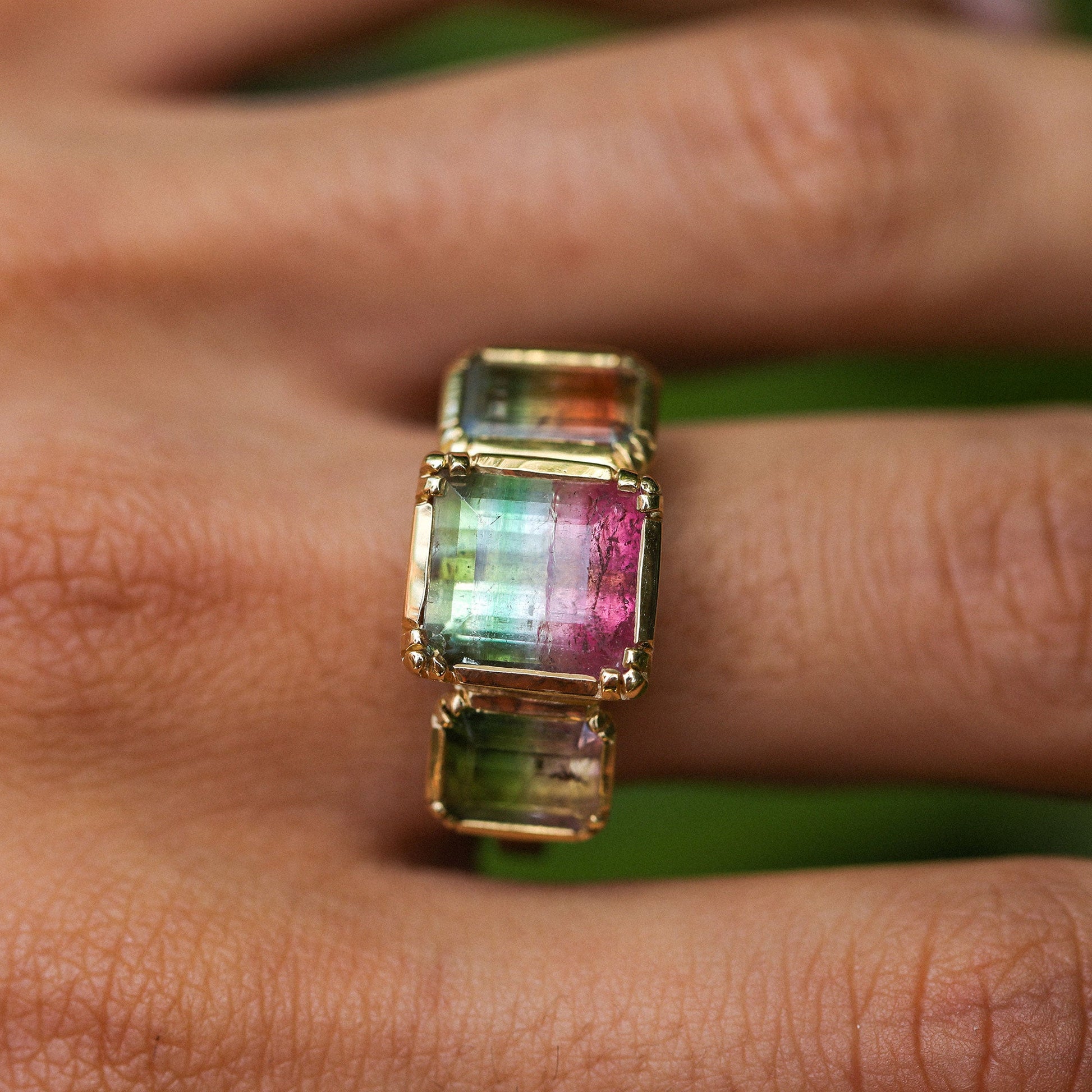 A gold ring with three rectangular iridescent bicolour tourmaline gemstones in green, pink, and blue tones, shown worn on a person's finger.