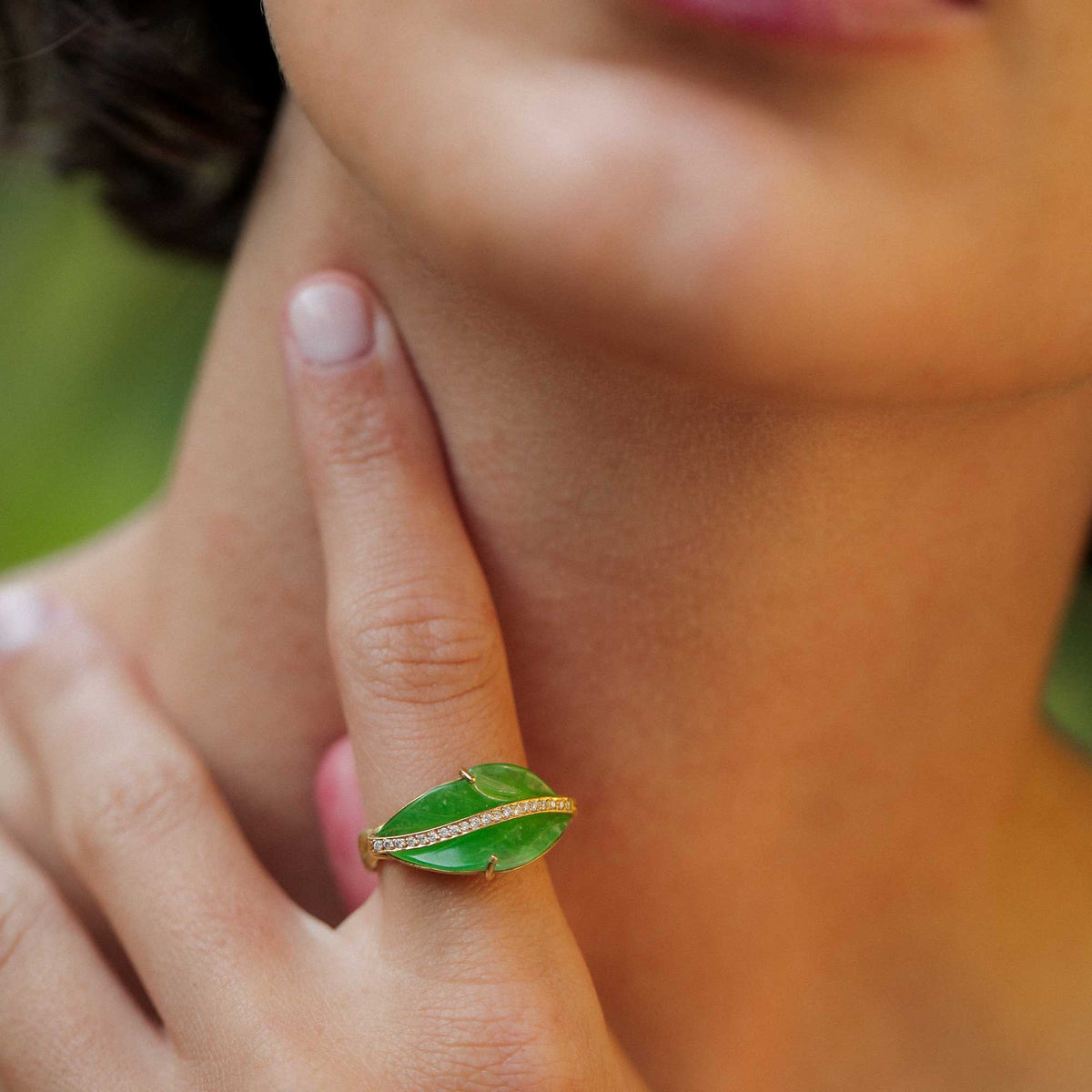 A close-up lifestyle shot of a person wearing a green jade leaf-shaped ring with a gold band studded with small diamond stones on their index finger.
