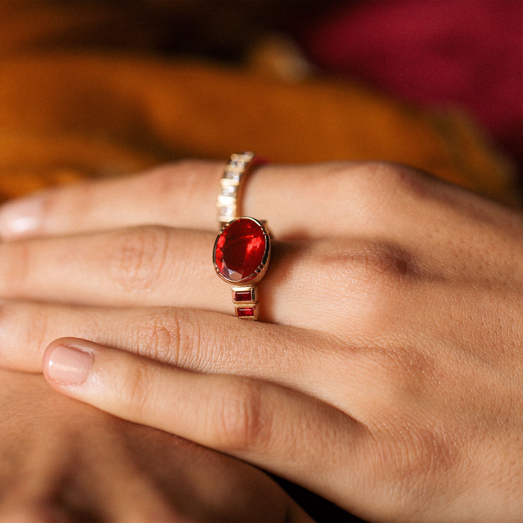 Close-up of a hand wearing a 18k gold ring with a red fire opal gemstone on a blurred background