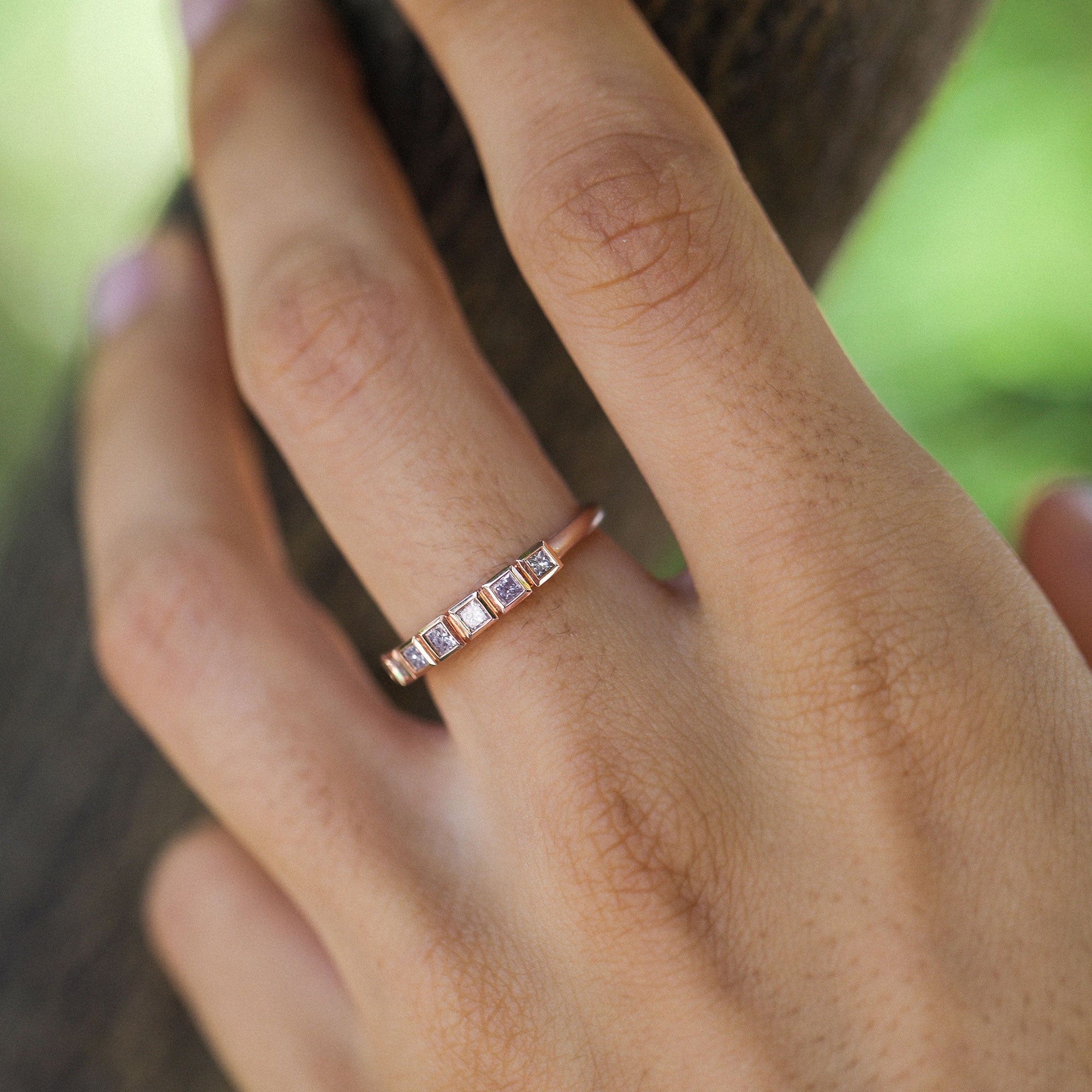 A close-up of a hand wearing a delicate rose gold band ring with five small square-cut pink diamond stones set in a row.