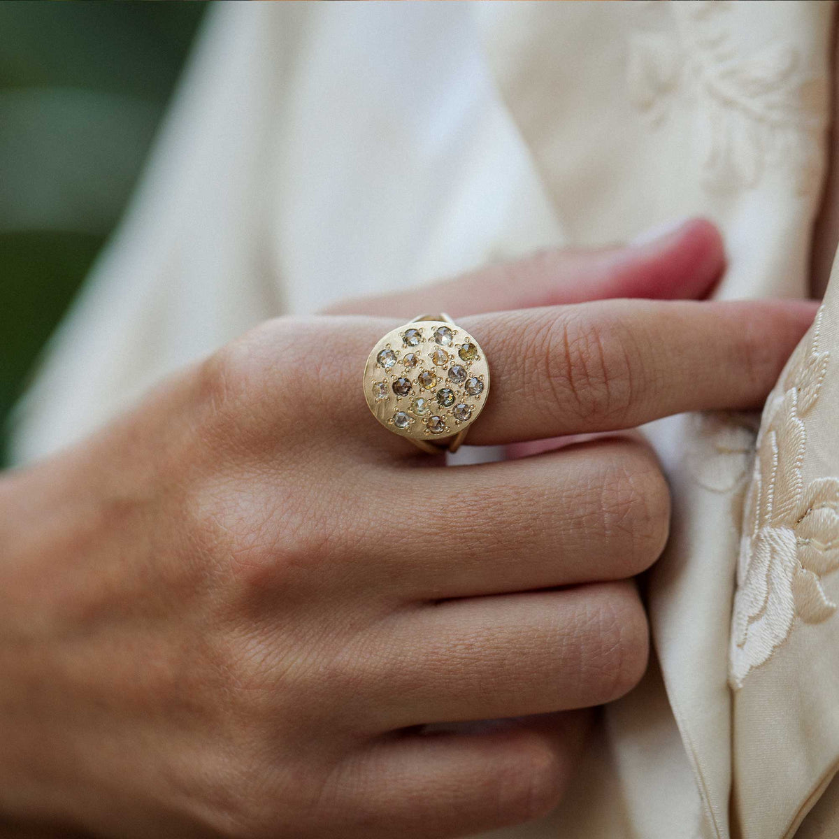 A close-up of a hand wearing a gold ring with a round face set with small clear and brown diamond gemstones, styled with cream-colored embroidered clothing.