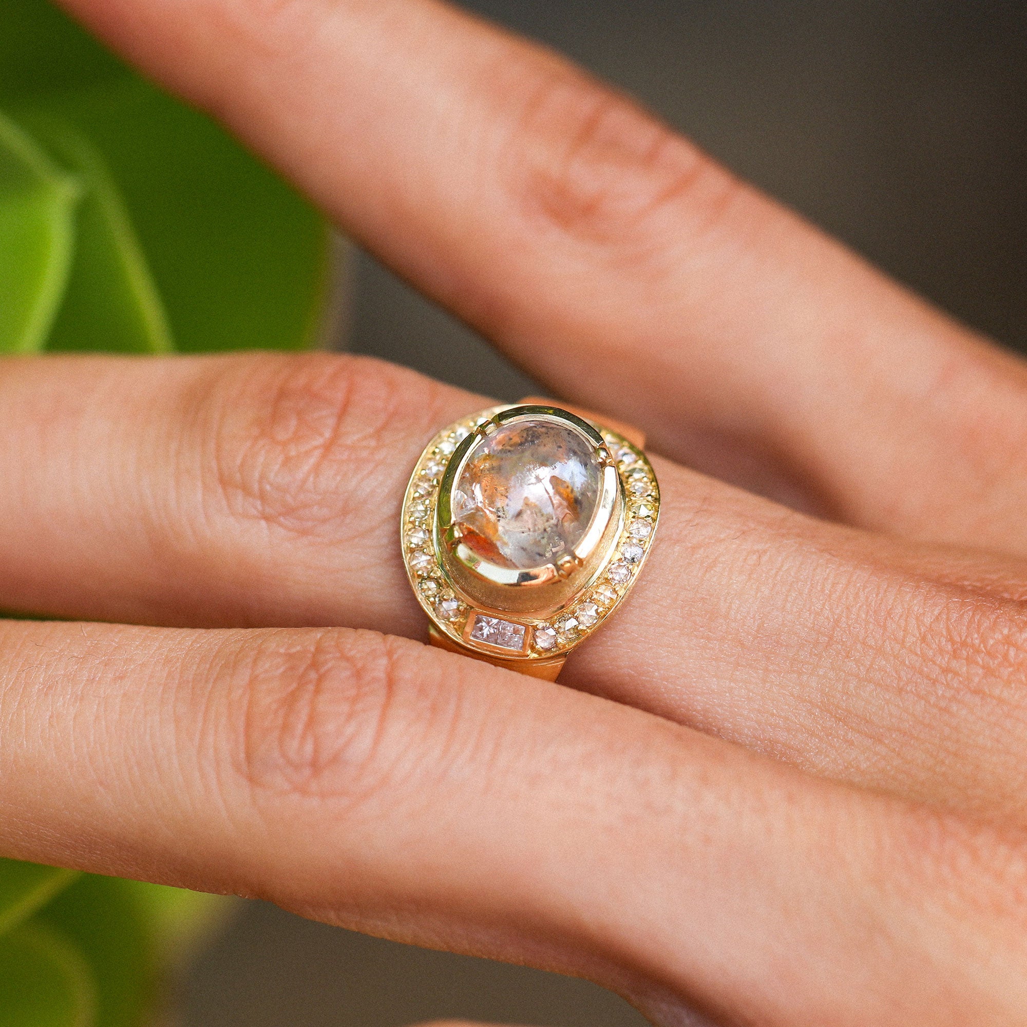 A gold ring with a large oval raw diamond gemstone and a halo of small diamond stones, shown worn on a person's finger against a soft blurred background.