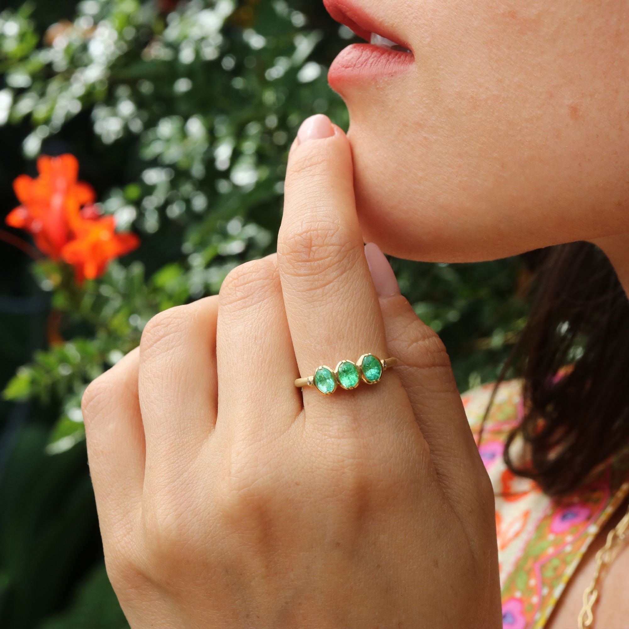 Hand wearing a gold ring with green paraiba tourmalines gemstones against a blurred natural background