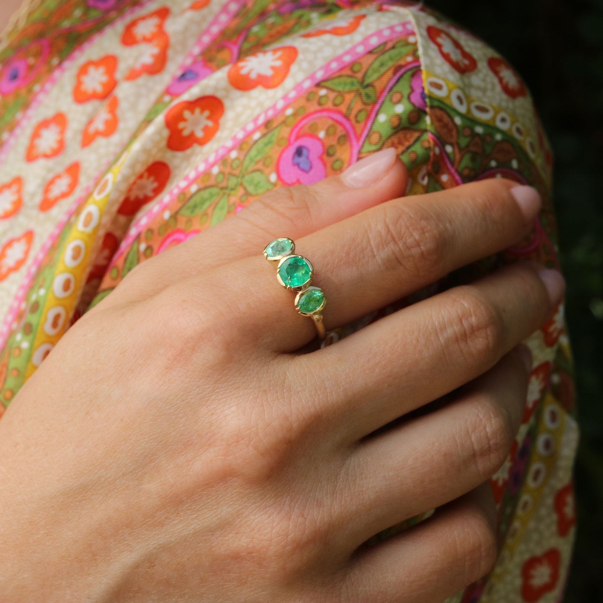 Hand wearing a ring with three green emerald and paraiba gemstones against a colorful fabric background