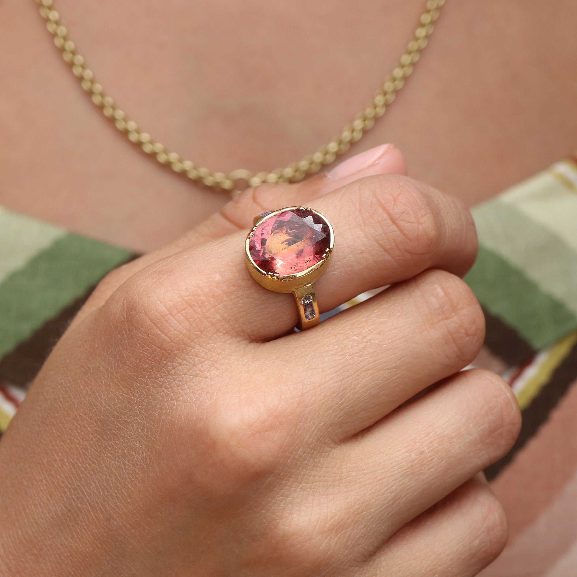 A gold ring with a large oval pink tourmaline gemstone, worn on a person's finger, with a gold chain necklace visible in the background.