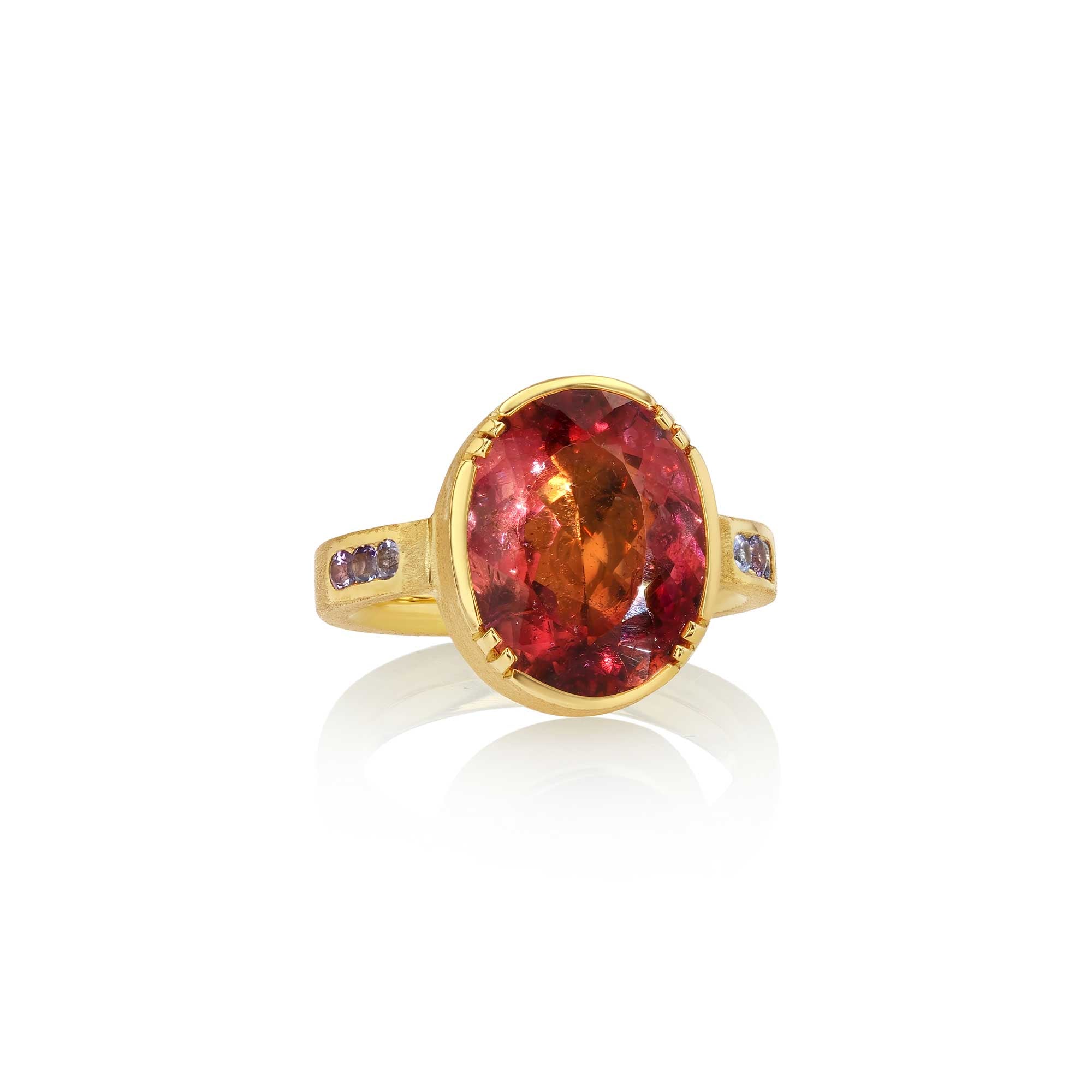 A yellow gold ring featuring a large oval-cut red tourmaline center stone, accented by small dark purple tanzanite stones on the shoulders, set against a clean white background with a soft reflection.