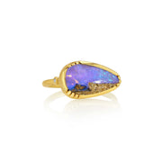 A gold ring featuring a large, irregularly shaped teardrop-shaped wood opal stone with vibrant blue, purple, and orange iridescent colors, accented by a small diamond gemstone on the band, set against a clean white background with a soft reflection.
