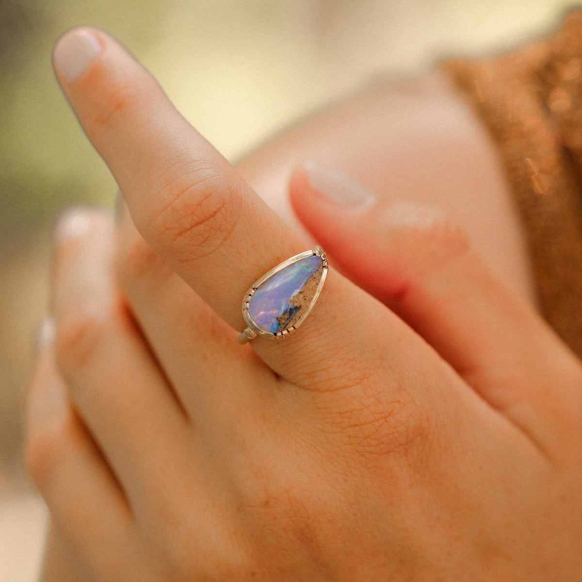 A close-up of a hand wearing a gold ring with a teardrop-shaped iridescent fossilised wood opal gemstone that shifts between blue, purple, and green.