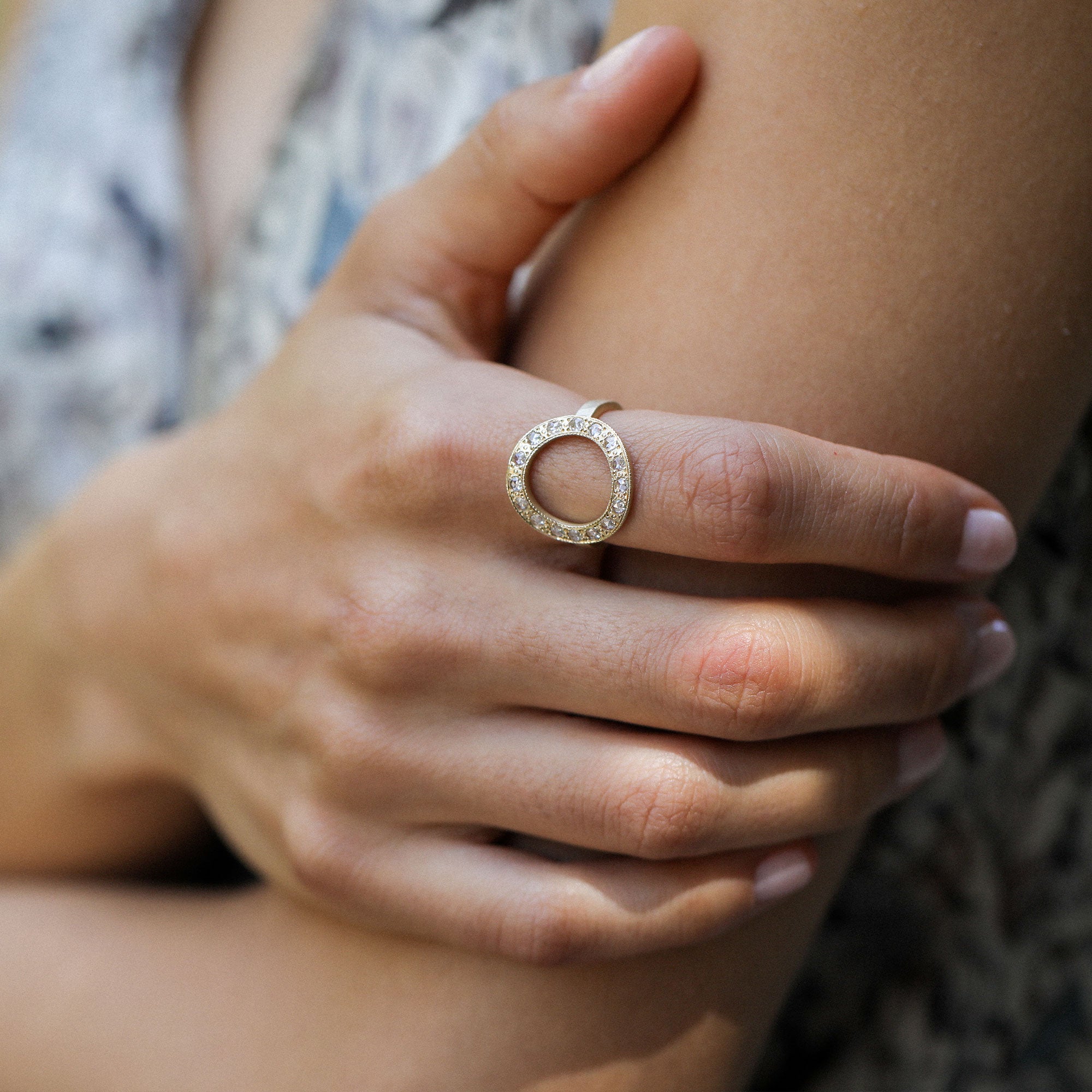 A close-up lifestyle shot of a gold ring with a halo setting of small clear diamond stones, worn on a person's finger.