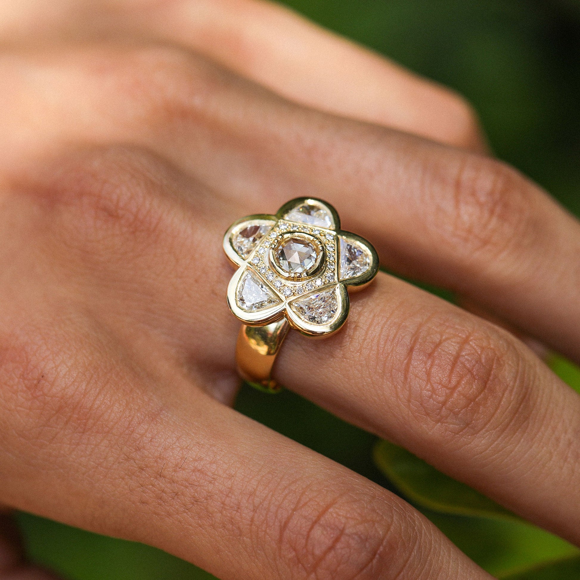 An 18k gold flower-shaped ring with diamond gemstones worn on a person's finger, set against a soft green blurred background.