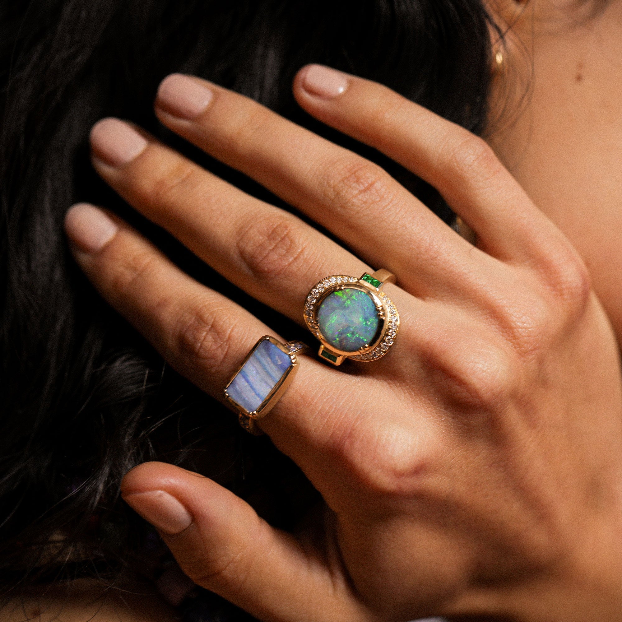 Close-up of a hand wearing two rings with gemstones, one blue and one green, against a blurred background.