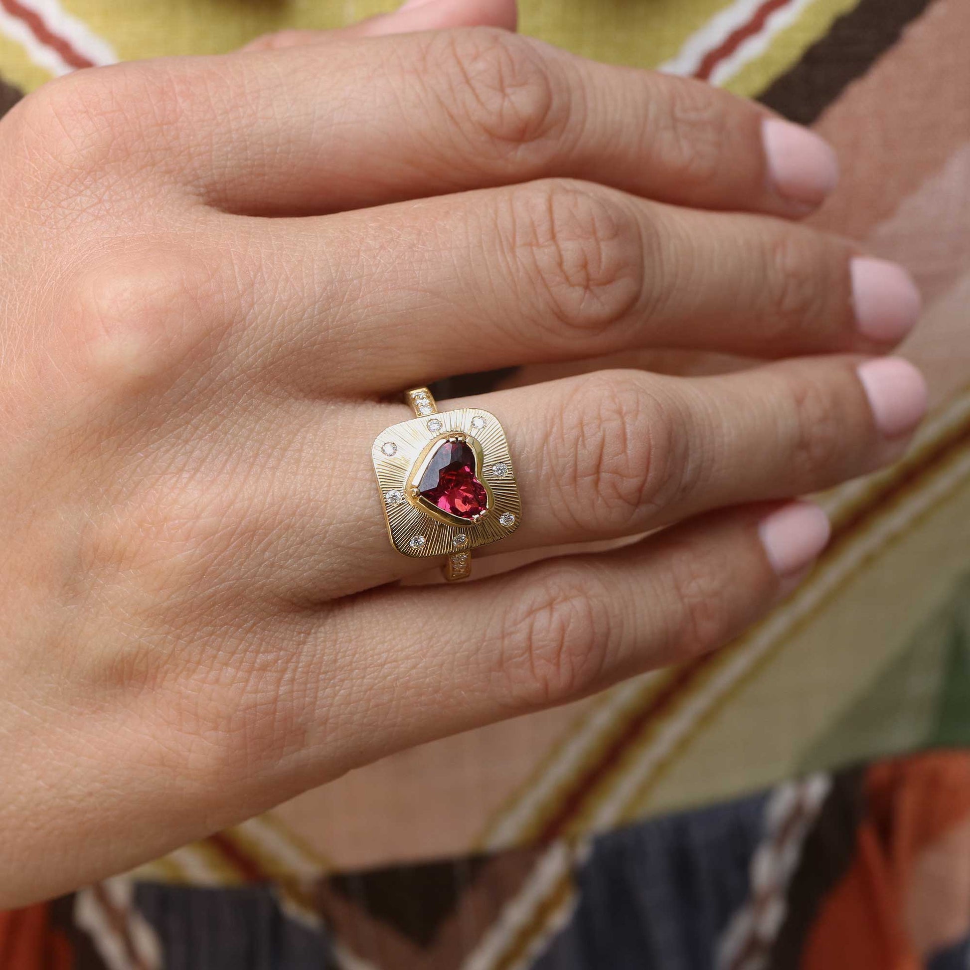 A gold ring with a heart-shaped red tourmaline gemstone and small clear stones, worn on a hand with pink manicured nails against a patterned background.
