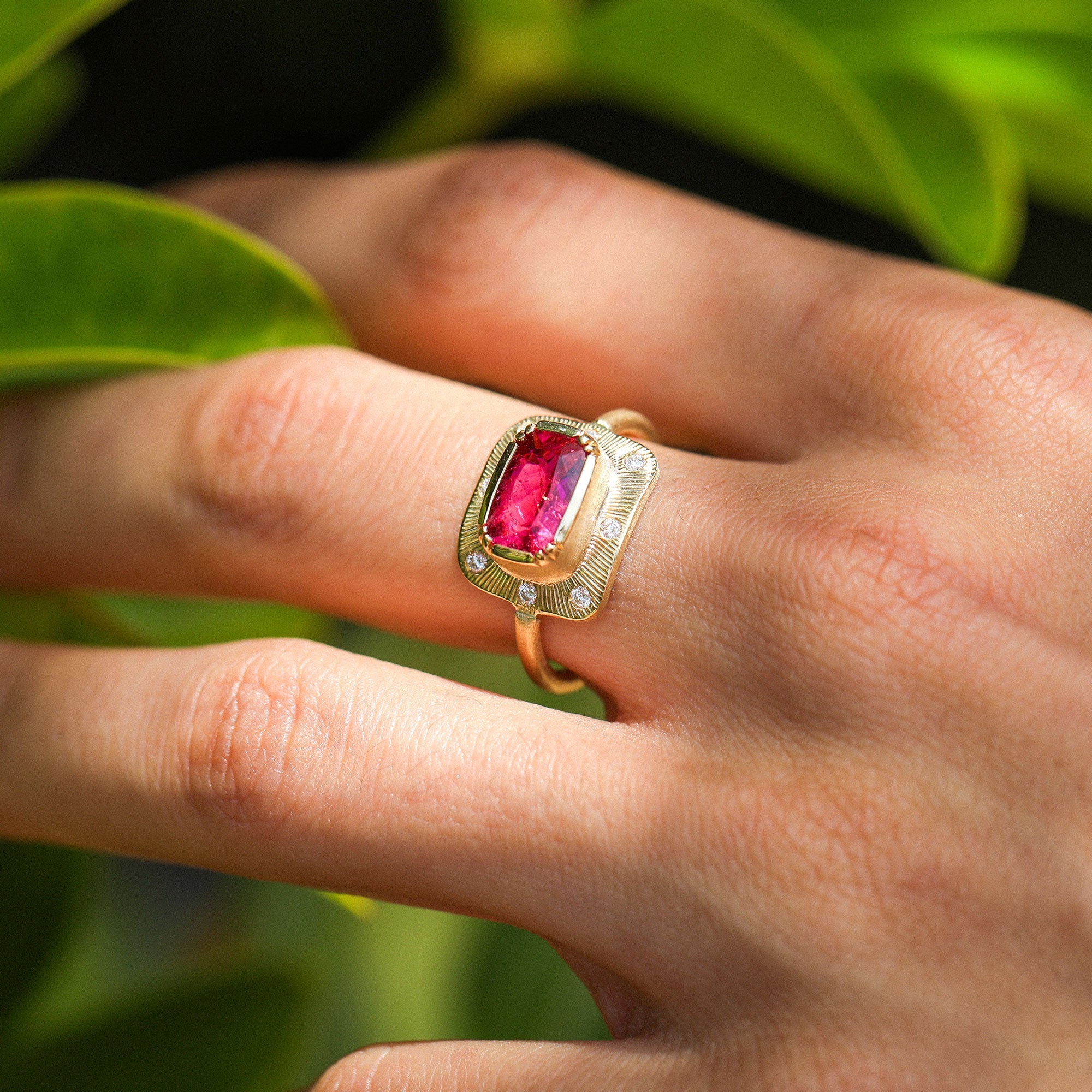 A close-up of a hand wearing a gold ring with a rectangular pink tourmaline gemstone and small diamond accent stones, set against a blurred green leaf background.