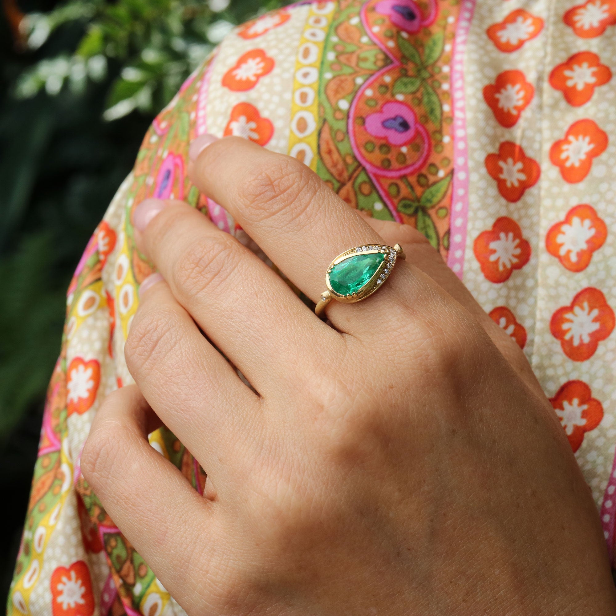 Hand wearing a green emerald ring with a colorful garment in the background