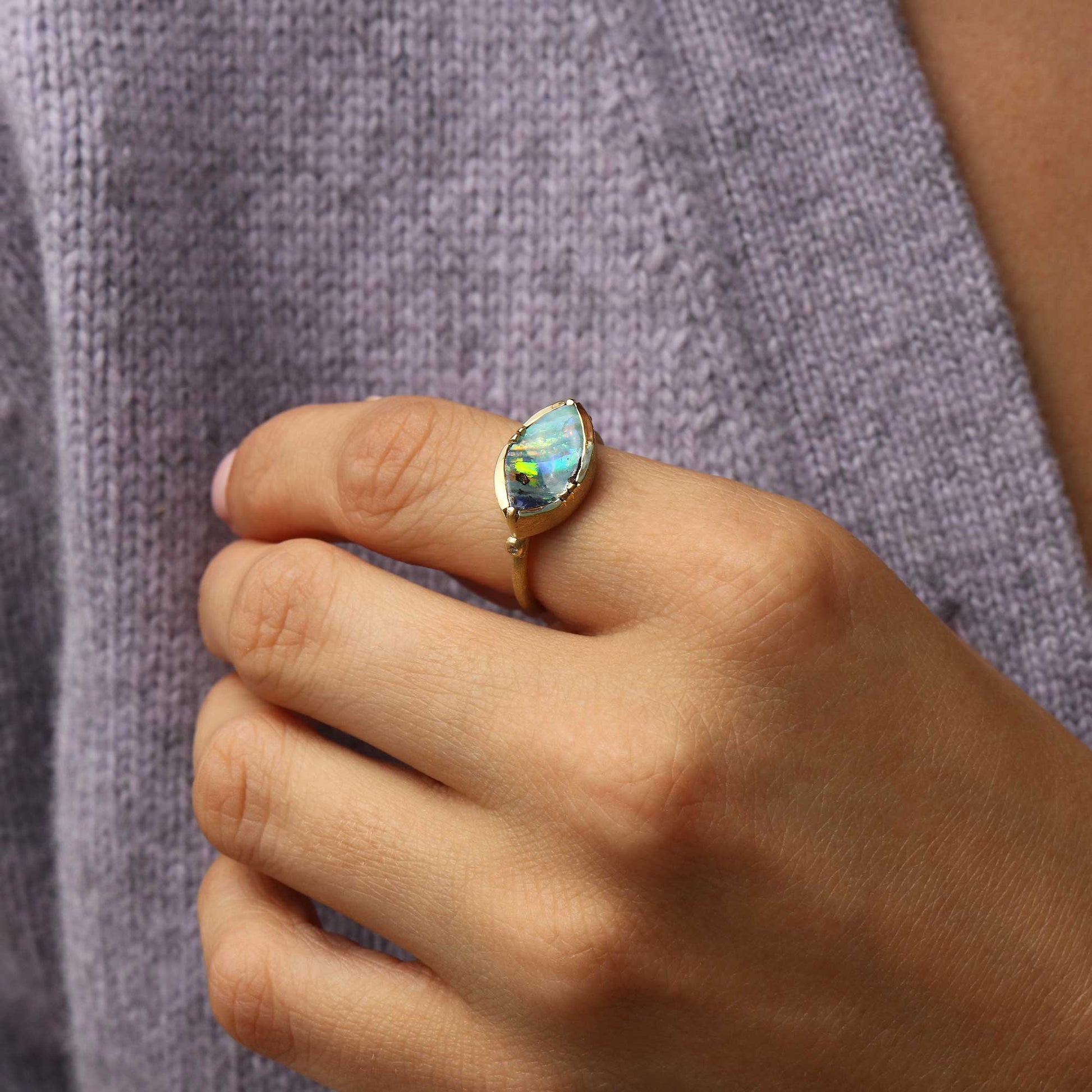 A close-up of a person's hand wearing a gold ring with a marquise-shaped iridescent blue-green boulder opal gemstone, set against a textured lavender knit sweater.