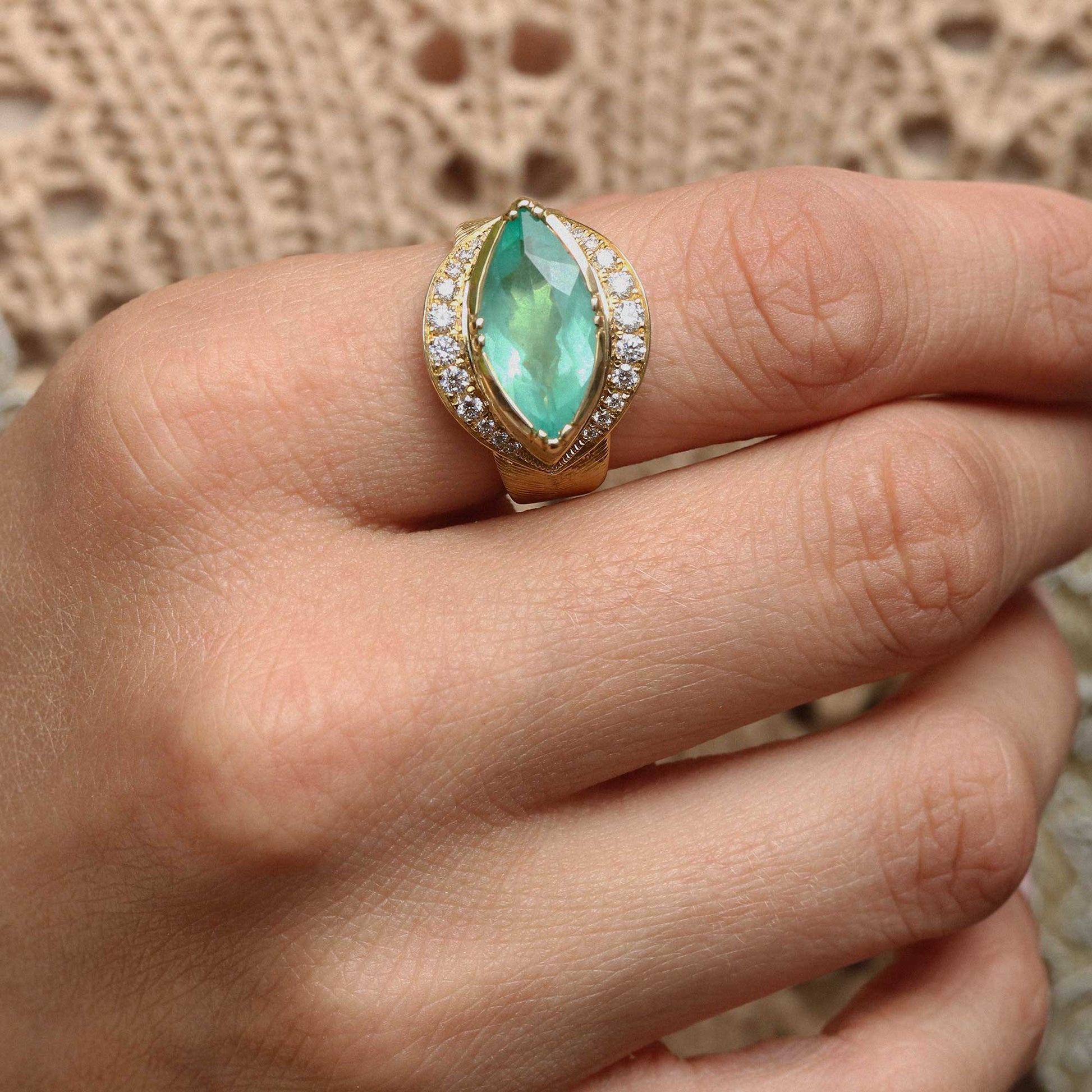 A close-up of a hand wearing a gold ring with a marquise-cut light green gemstone and small clear accent stones, set against a soft beige textured background.