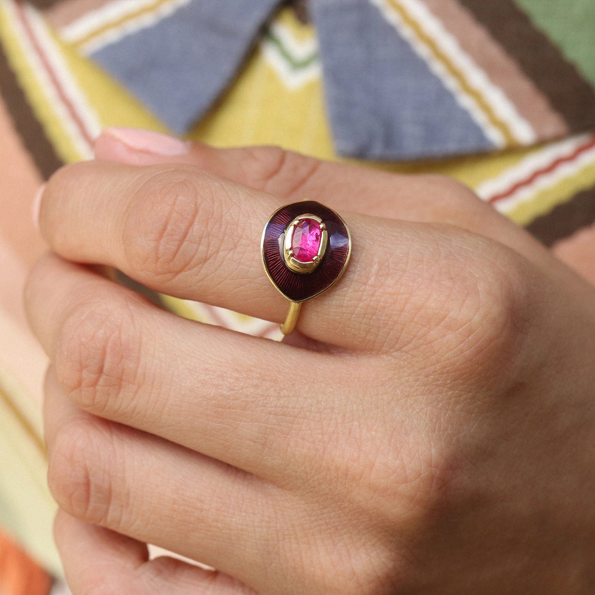 A gold ring with a pink oval gemstone and a dark purple enamel halo, worn on a person's finger against a colorful patterned background.