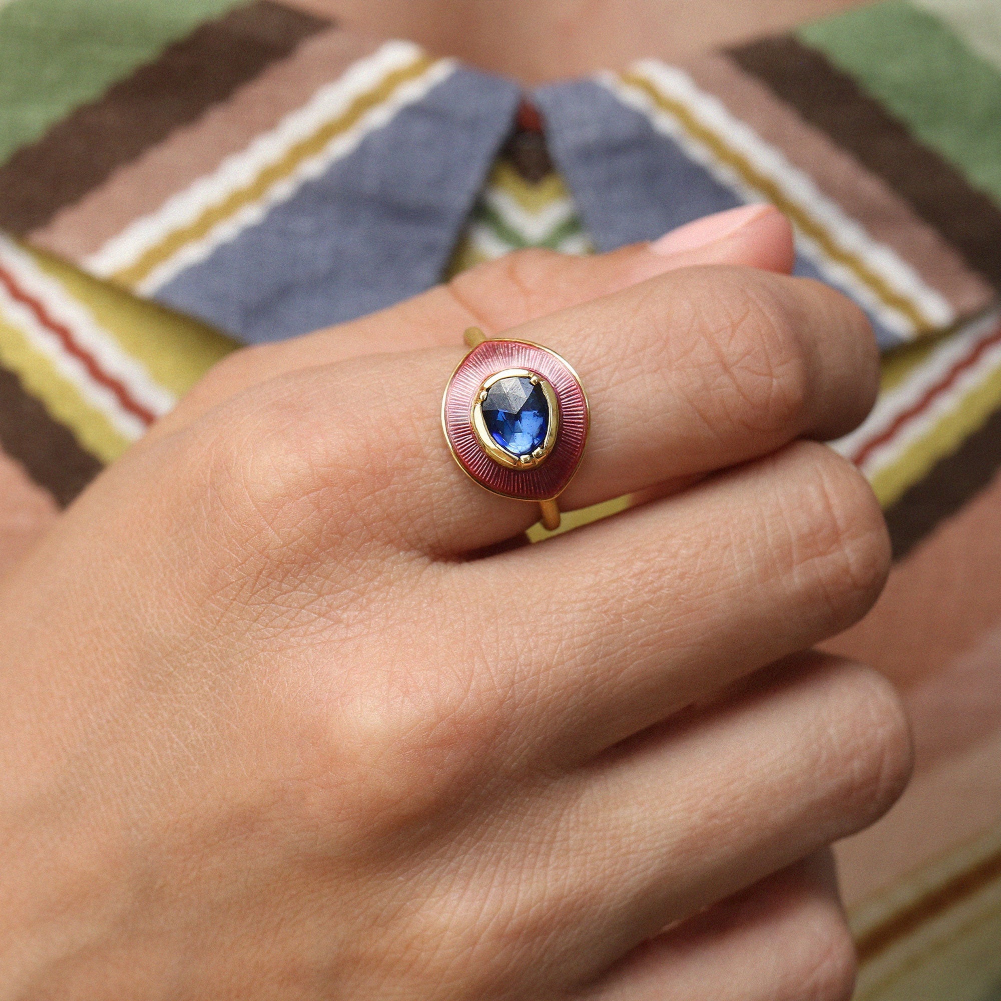 A gold ring featuring a central oval blue sapphire gemstone surrounded by a textured, lilac circular enamel setting, all on a polished gold band worn on a persons finger against a patterned colourful dress.