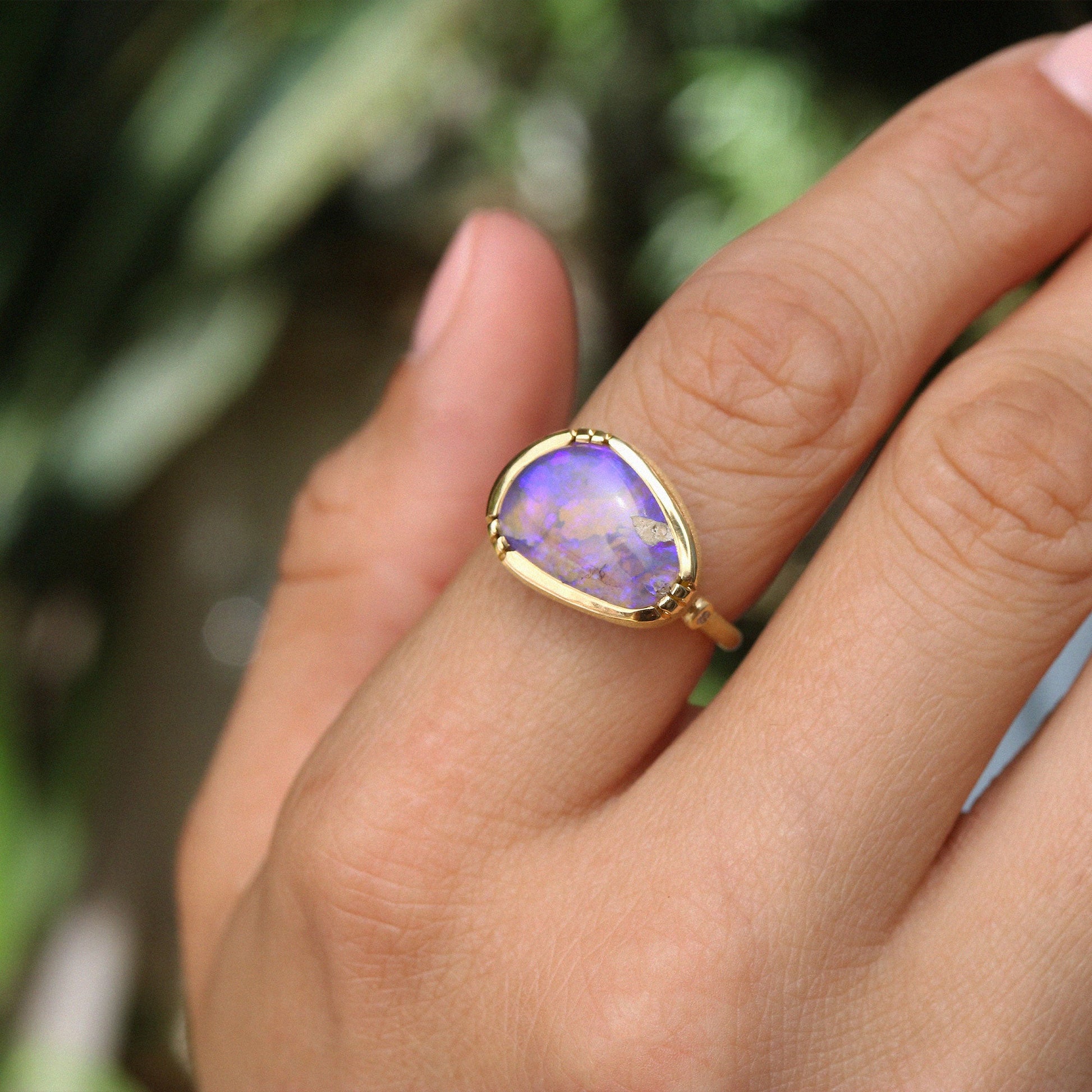 A gold ring with a teardrop-shaped purple opal gemstone, worn on a person's finger against a blurred green background.